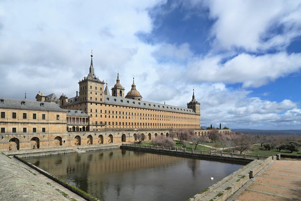Historic building with towers and arched facade, surrounded by a pond and landscaped gardens, under a partly cloudy sky.