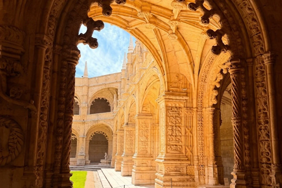 A historic cloister with ornate stone arches, intricate carvings, and a central grassy courtyard under a bright blue sky.