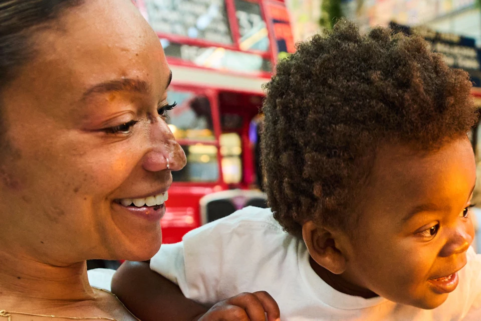 A smiling woman holding a child, who is reaching out excitedly. They are in an indoor space with red buses and people in the background.