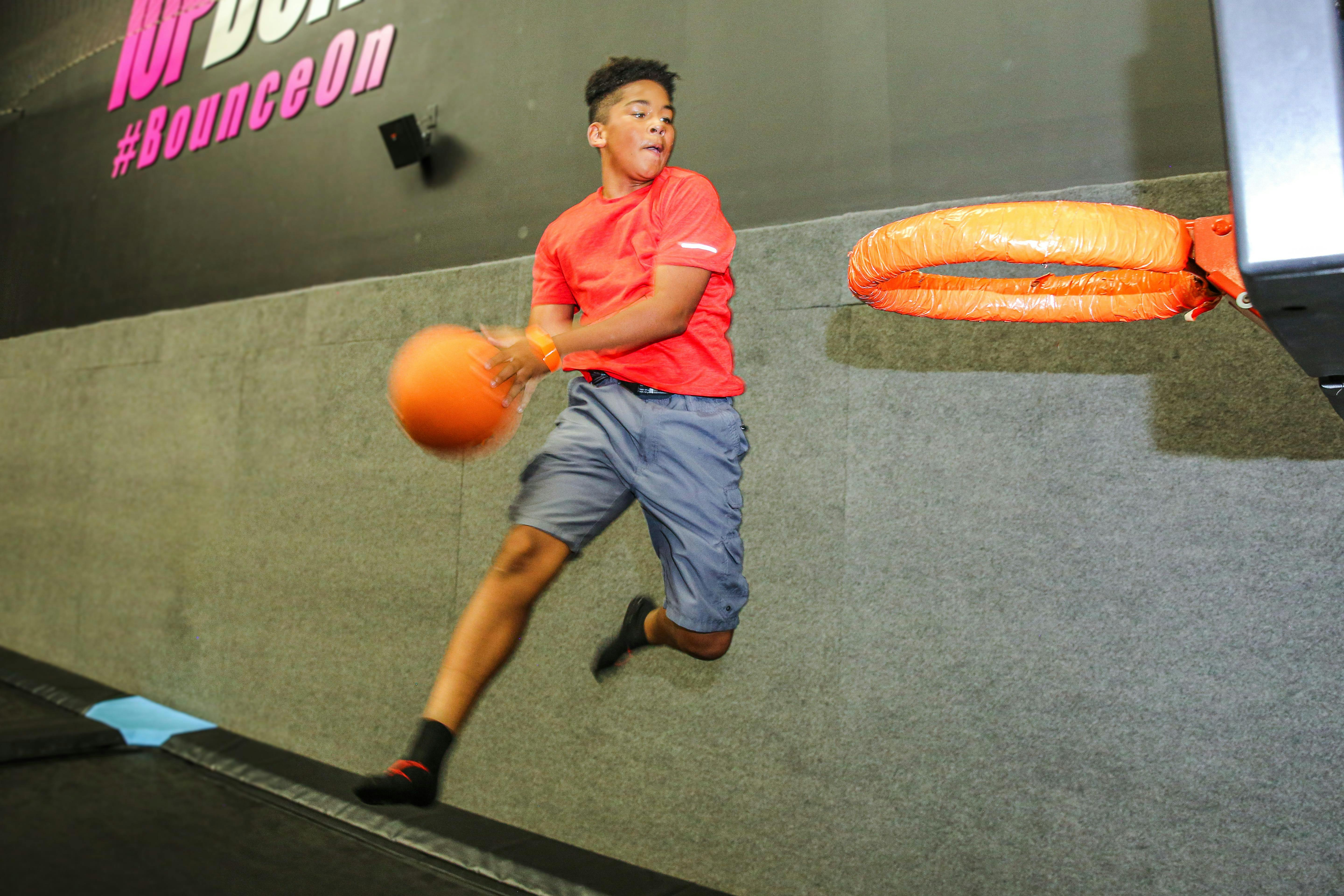 A boy in a red shirt and gray shorts is mid-air, about to dunk a basketball into an orange hoop, with a wall in the background.