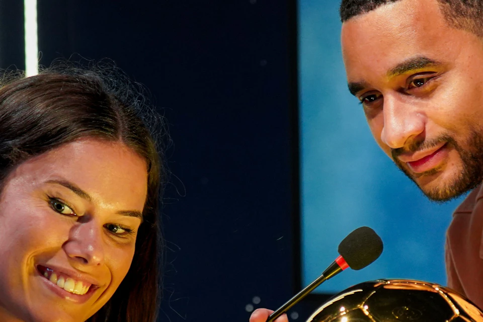 Two people are posing with a golden football trophy on a glass stand. One of them is pointing at the trophy whilst holding a mobile phone.
