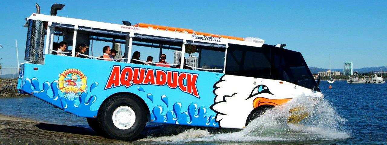 Kolorowy, amfibijny pojazd wycieczkowy z etykietą „Aquaduck” wjeżdżający do wody, z pasażerami na pokładzie, pod czystym, błękitnym niebem.