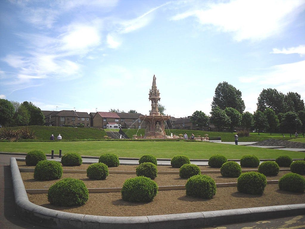 Un jardin bien entretenu avec une haie circulaire, une grande fontaine ornée et des gens qui se promènent et s&#39;assoient sur des bancs.