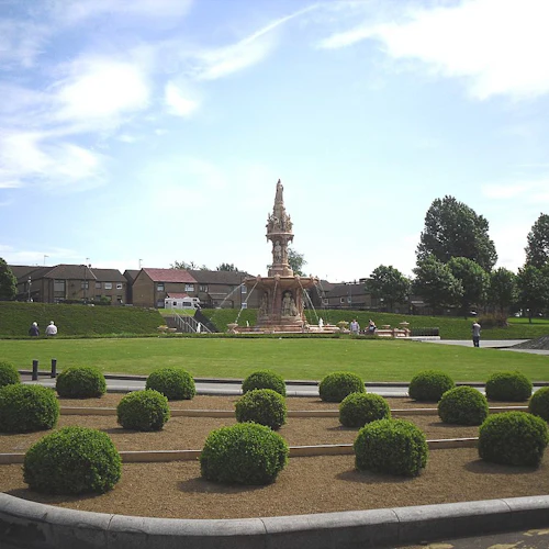 En välskött trädgård med ett cirkulärt häckarrangemang, en stor utsmyckad fontän och människor som går och sitter på bänkar.