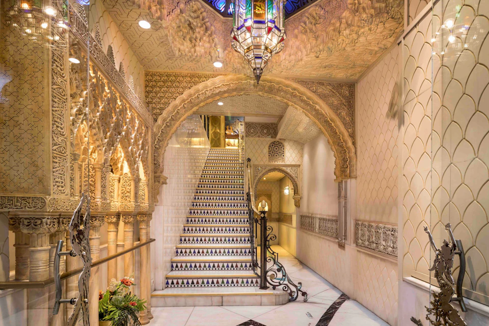 Ornate interior with intricate arches, a staircase with patterned tiles, a hanging chandelier, and decorative details.