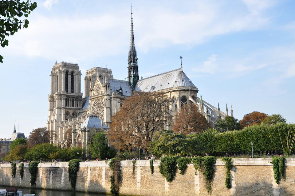 A Gothic cathedral with ornate architecture seen from across a river, surrounded by trees under a clear sky.