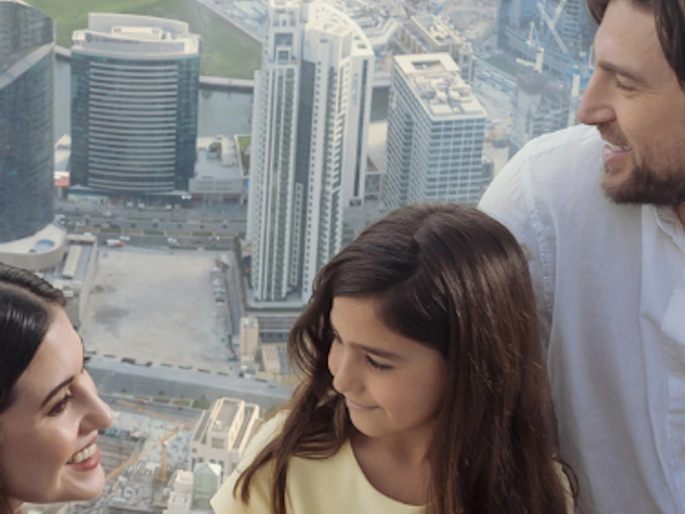 A man, woman, and young girl sit together on a high observation deck overlooking a large city skyline with tall buildings.