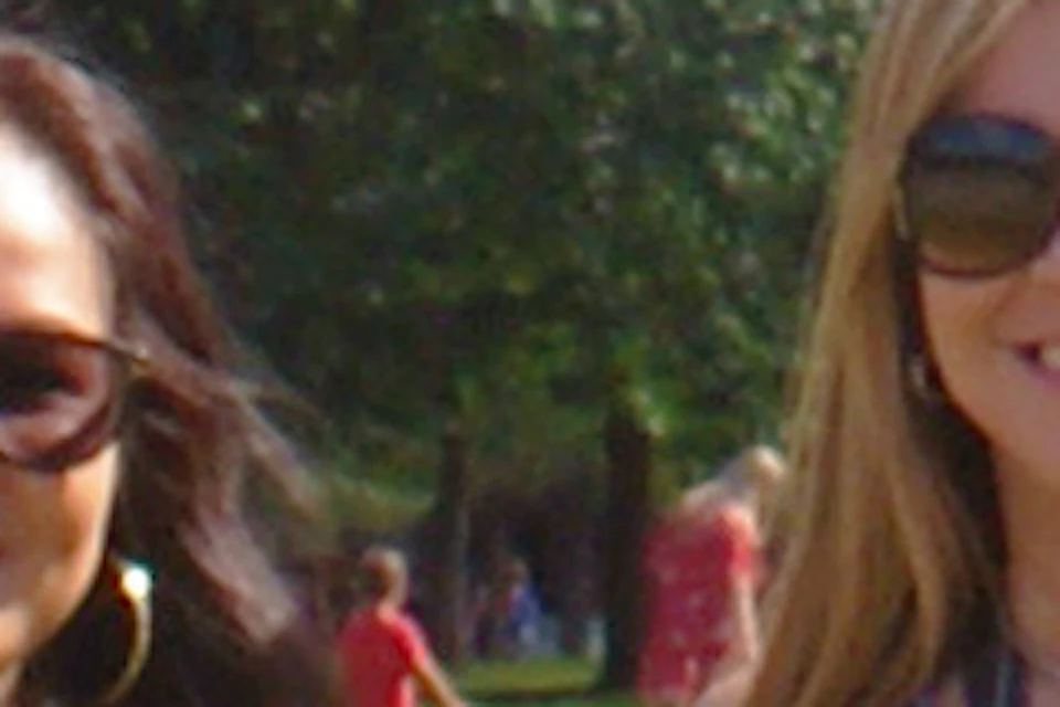 Two women pose for a photo in a park with a historic building and skyscrapers in the background, while another person takes their picture.