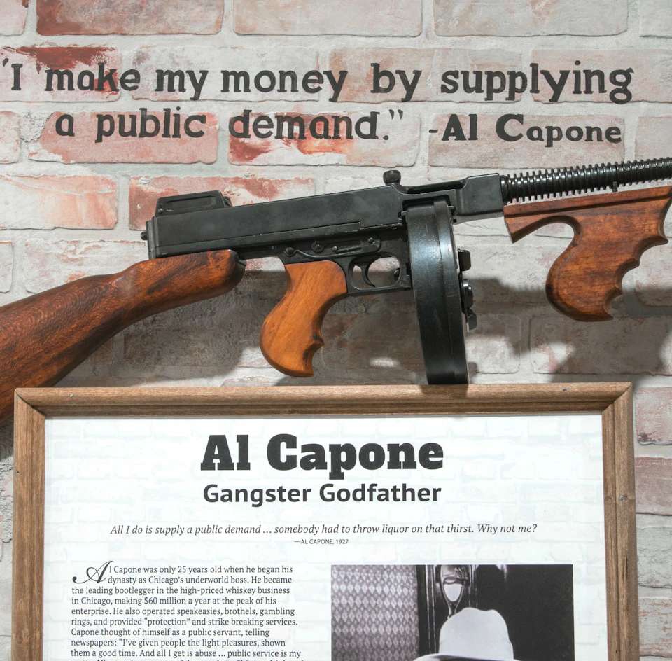 A framed document about Al Capone is displayed under a mounted submachine gun and a quote attributed to him on a brick wall.