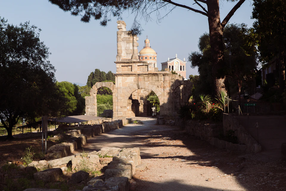 Ancient stone archway with a pathway leading through it. Two domed buildings in the background, surrounded by trees and greenery.
