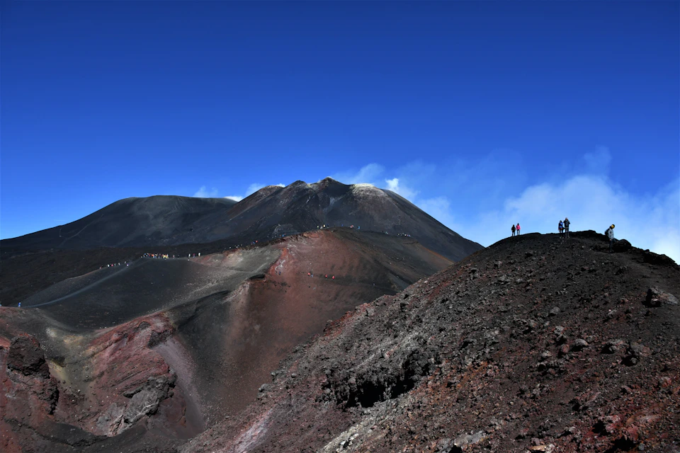 People hiking on a rugged, volcanic landscape with a mountain peak emitting light smoke under a clear blue sky.