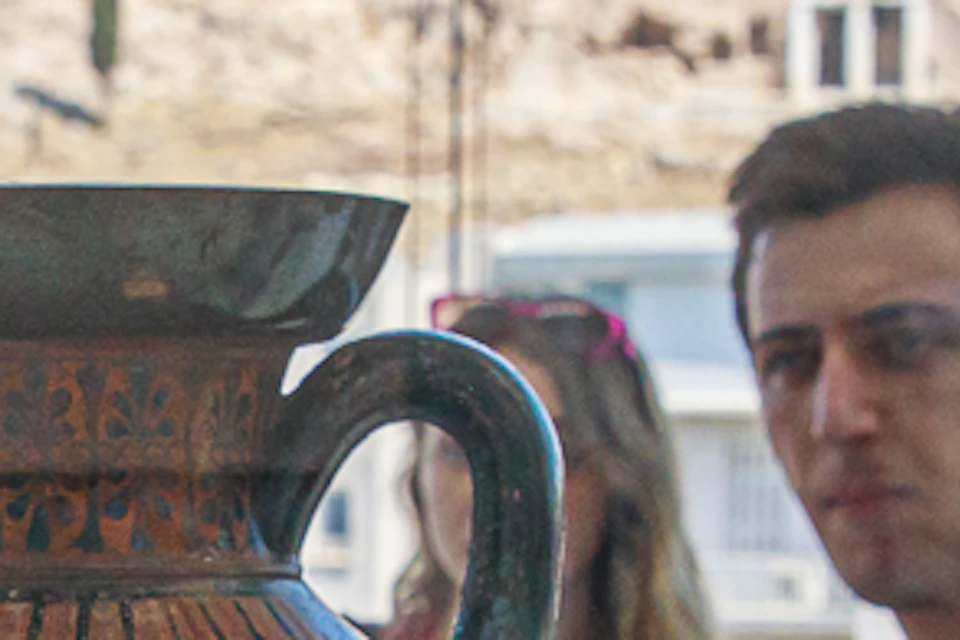 People view an ancient Greek vase with a racing scene in a glass case. The Acropolis is visible through the window behind them.
