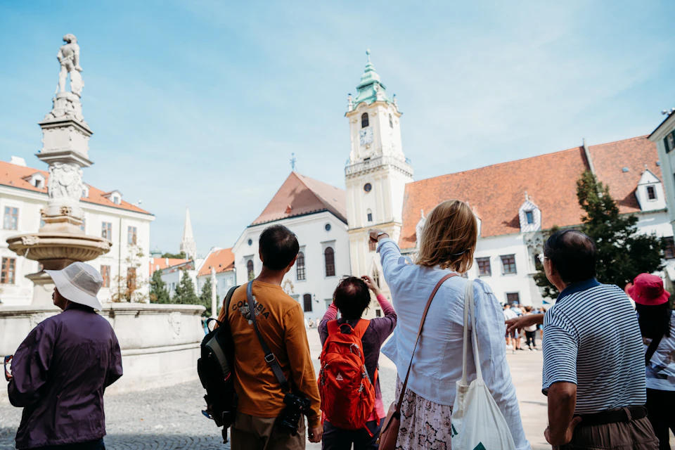 A group of people, including a person pointing, stand in a courtyard facing a white church with a tall, ornate tower and red roof.