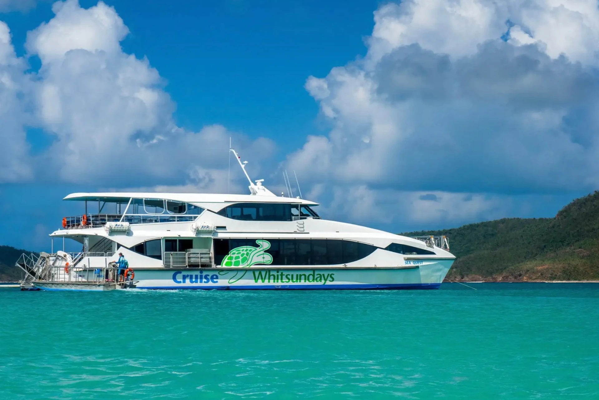 A white boat with "Cruise Whitsundays" and a turtle logo is on turquoise water under a partially cloudy blue sky.