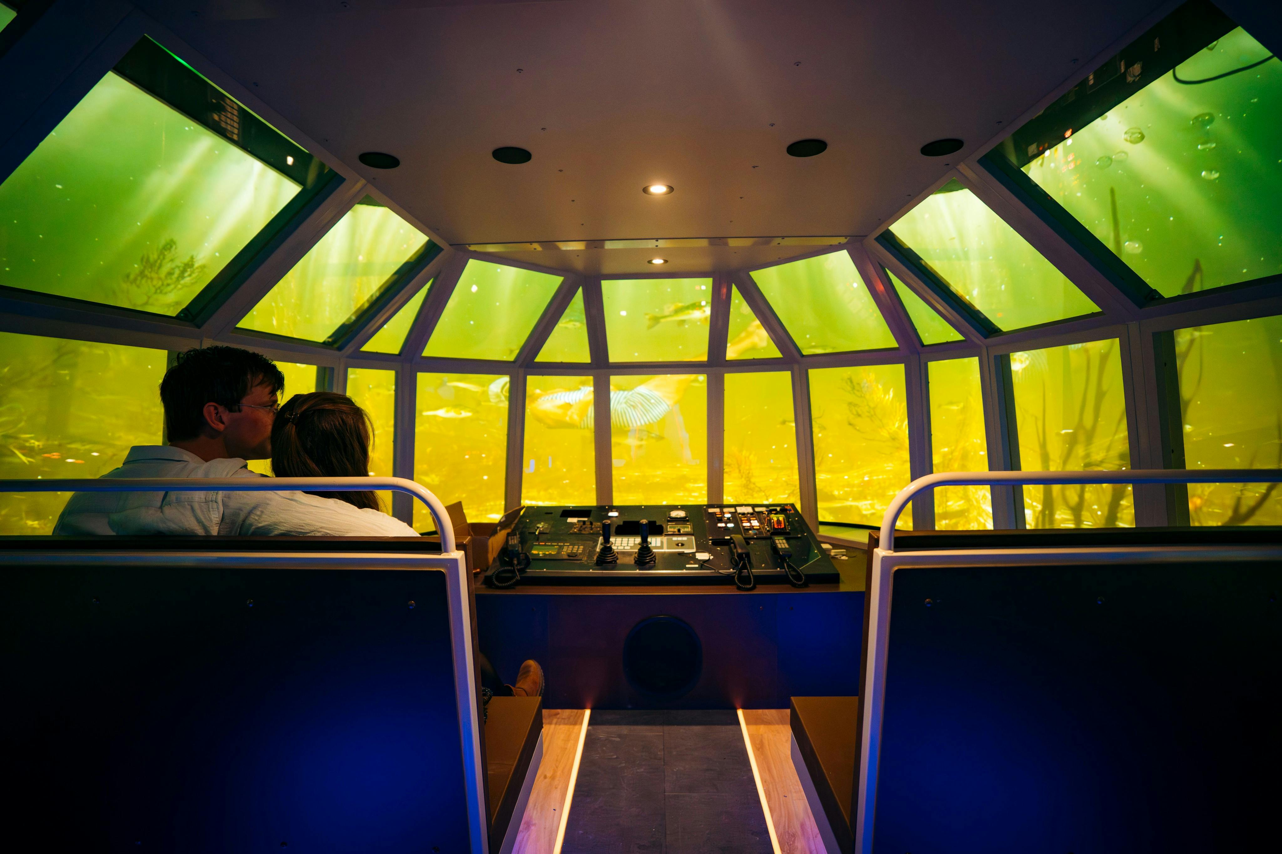 Två personer sitter i ett undervattensobservationsrum med en kontrollpanel, omgivet av stora fönster som visar en gult upplyst havsscen.