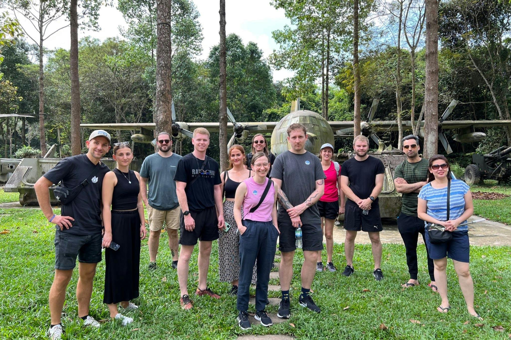 A group of people stands on a grassy area with trees and an old military airplane in the background.