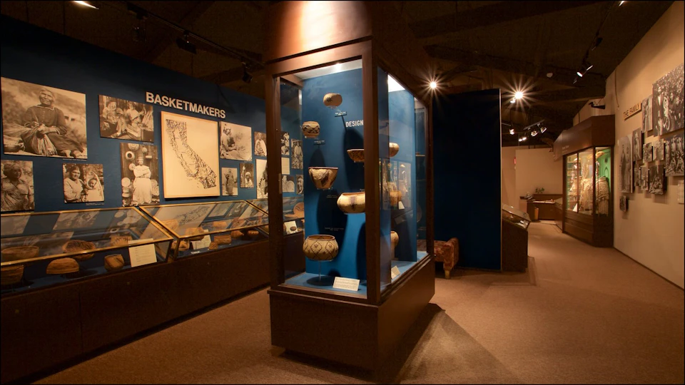 Museum exhibit displaying Native American baskets in glass cases, with wall posters and maps labeled "Basketmakers" and "Design."