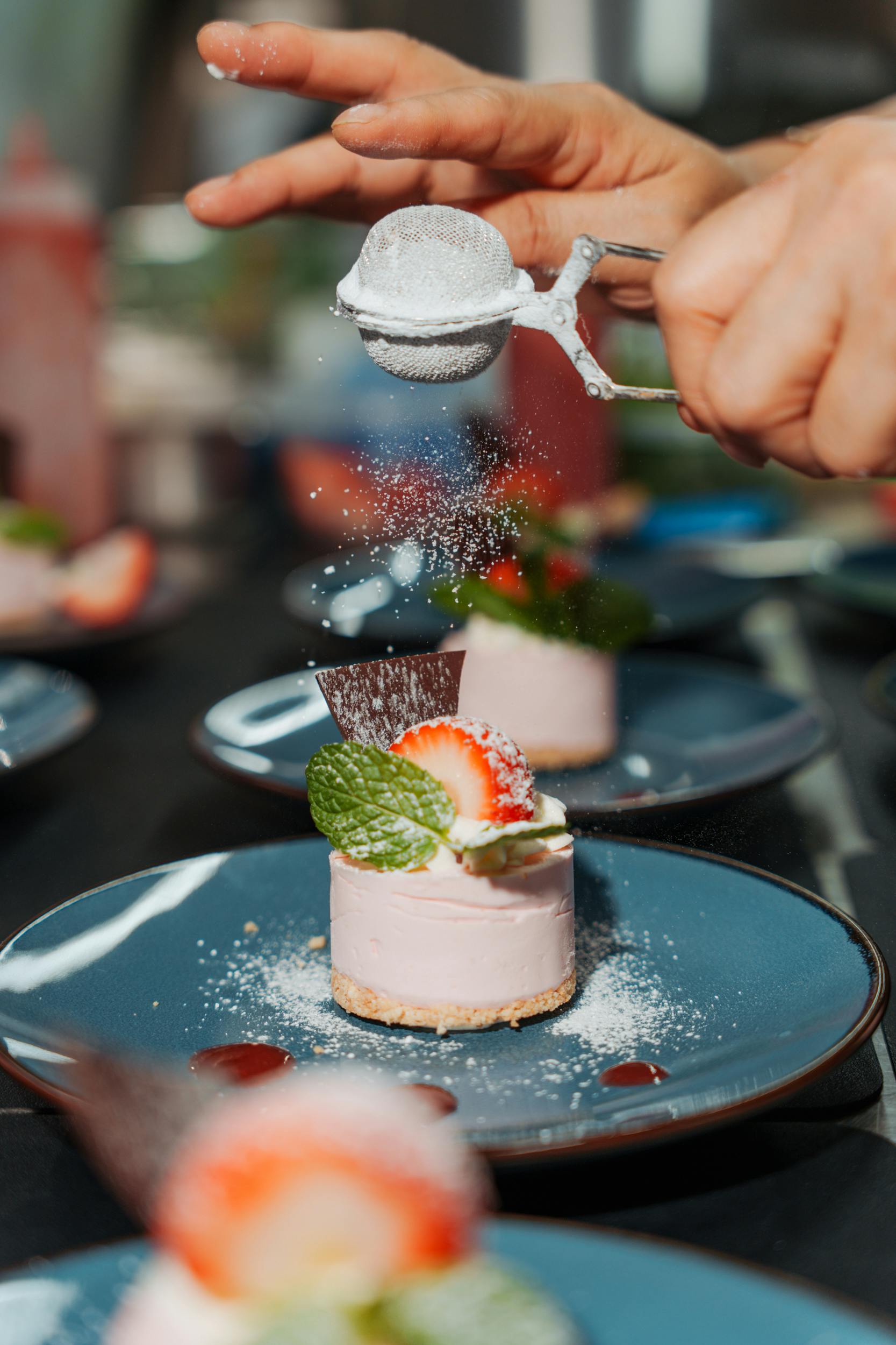 Een hand strooit poedersuiker op een aardbeien cheesecake gegarneerd met muntblaadjes op een blauw bord.
