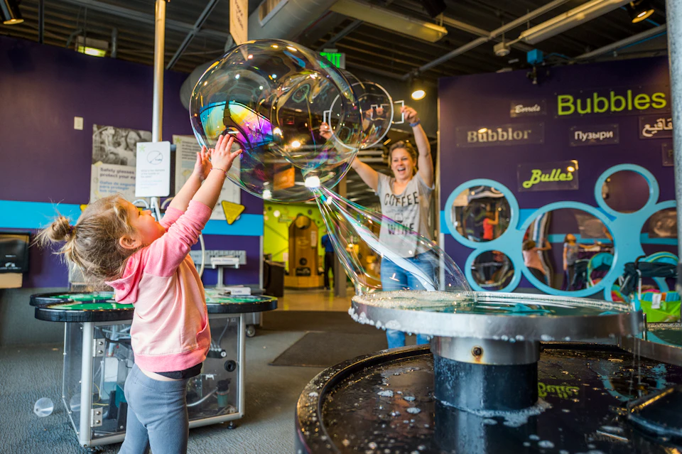 Children play with large, colorful bubbles at an indoor science museum exhibit. A woman in the background cheers excitedly.