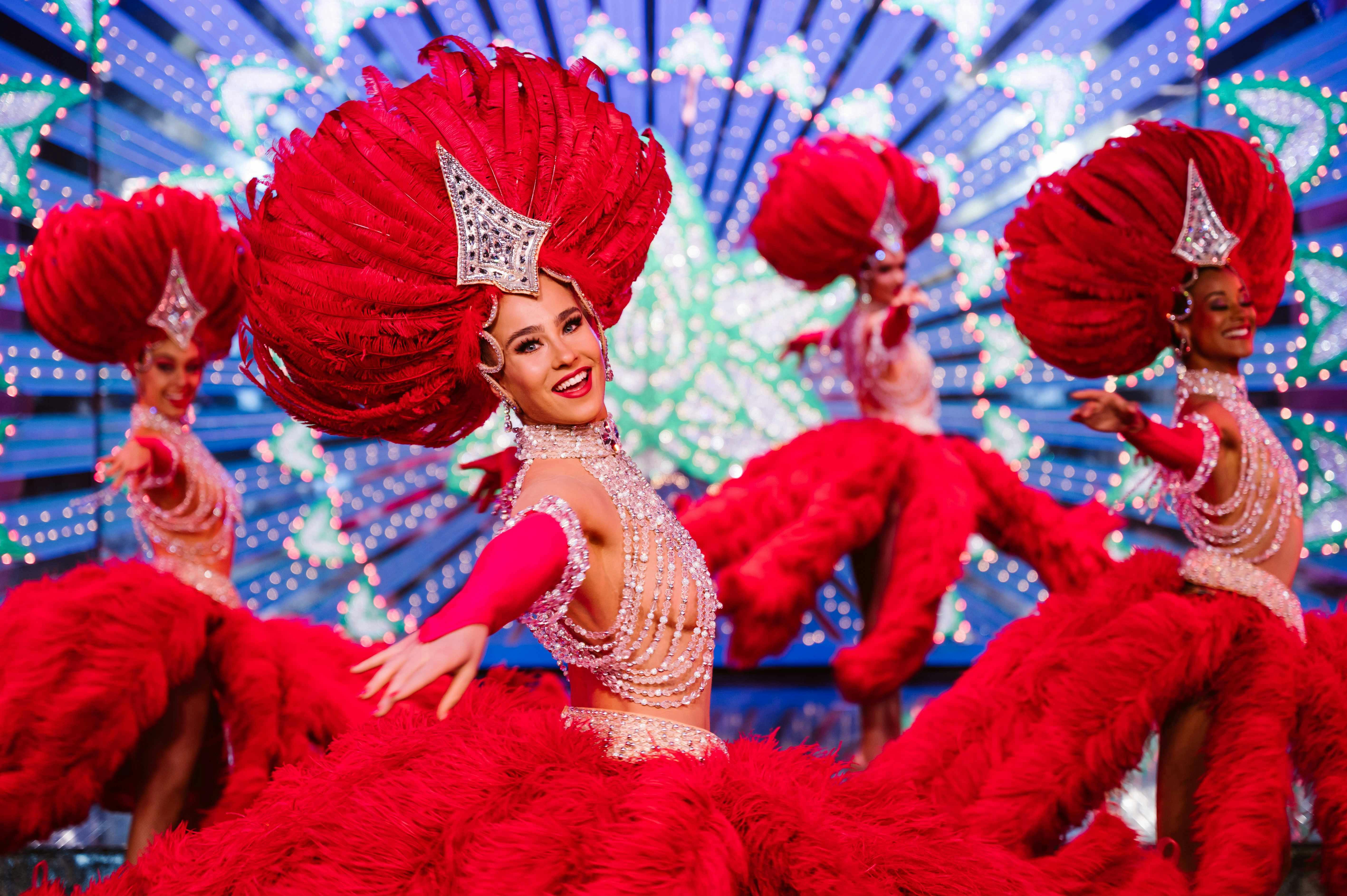 Performers in glamorous costumes with large red feathered headdresses and sequined outfits dancing on a vibrant stage.