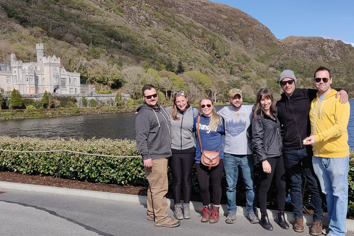 여섯 명이 카메라를 보며 웃으며 서로 가까이 서 있습니다. 산과 호수를 배경으로 야외 활동을 즐길 수 있습니다.