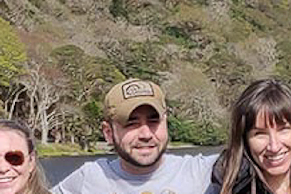 Six people stand closely together, smiling at the camera. They are outdoors with mountains and a lake in the background.