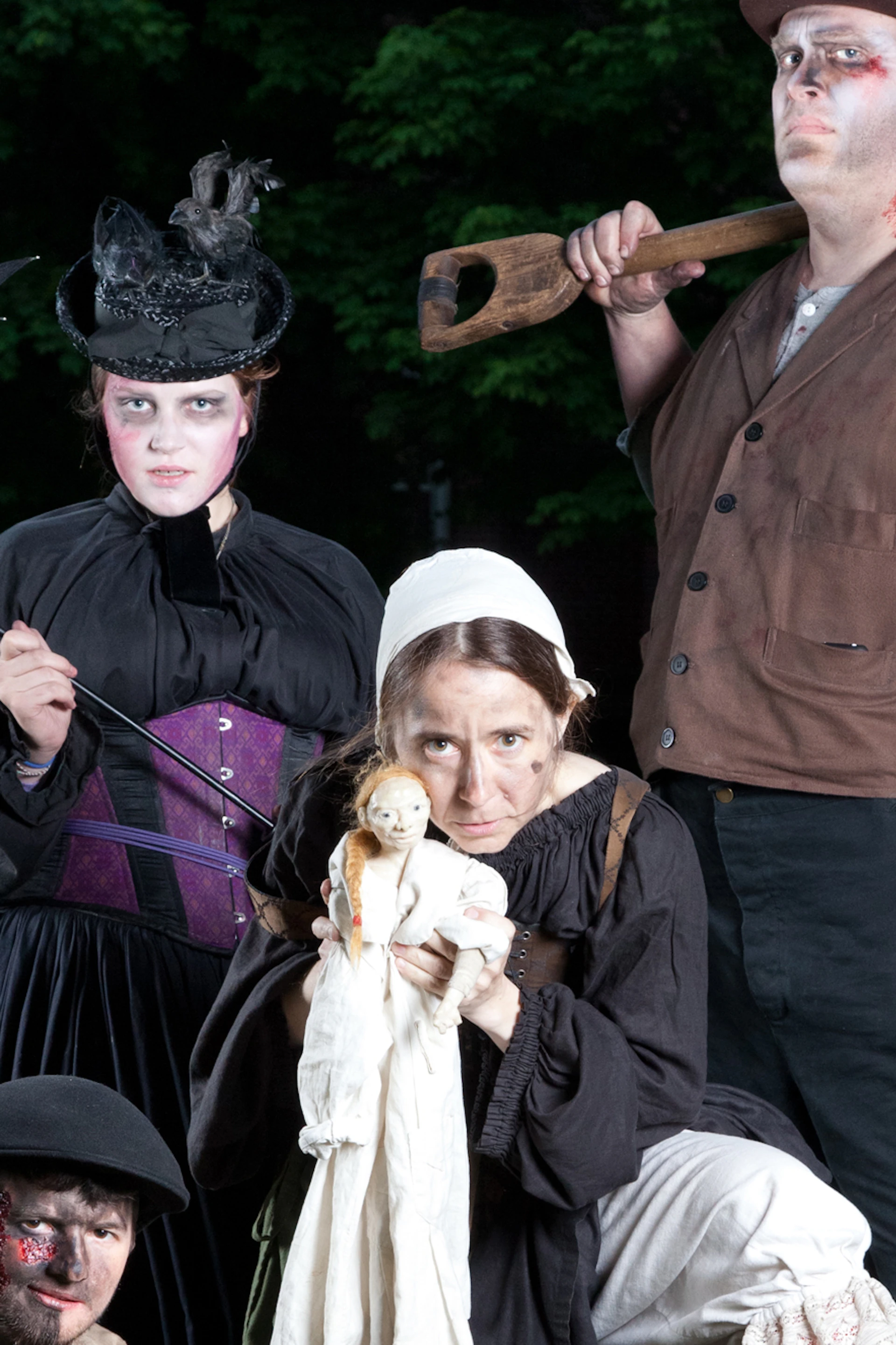 Four people in Victorian-style costumes with makeup, posing in a graveyard. One holds a shovel and another holds a doll.