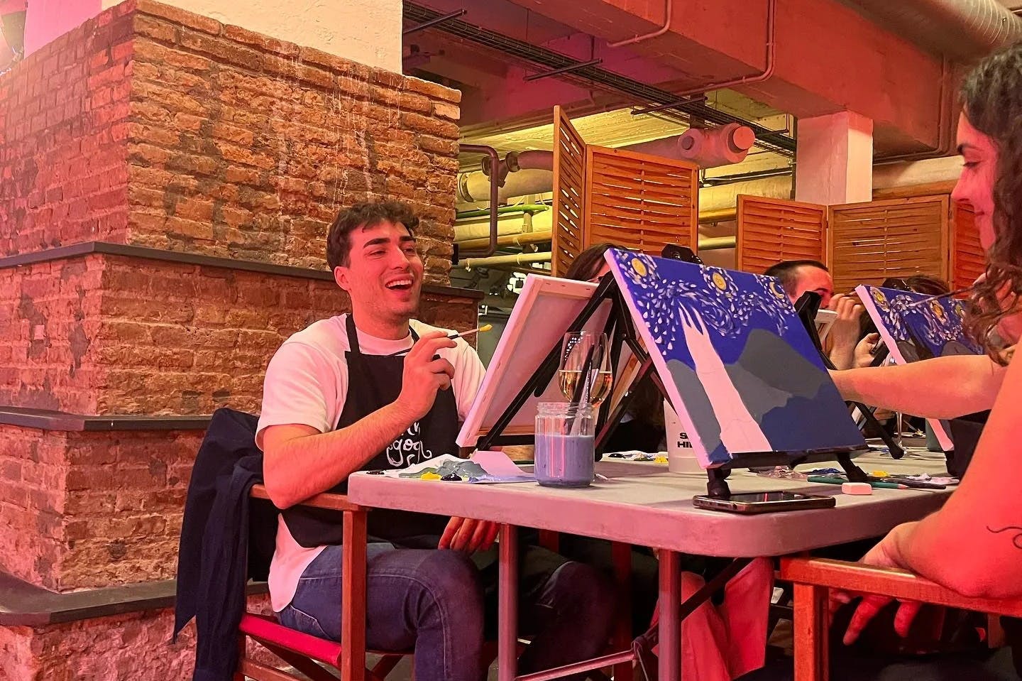 Ένα άτομο που φοράει λευκό πουκάμισο και ποδιά χαμογελά ενώ ζωγραφίζει ένα καβαλέτο μέσα σε έναν χώρο βιομηχανικού στιλ.