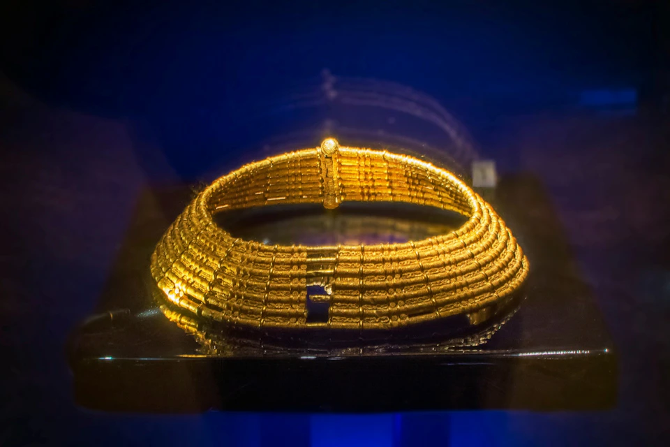 A golden, ancient-style collar made of multiple rows of intricate beads, displayed against a dark blue background.