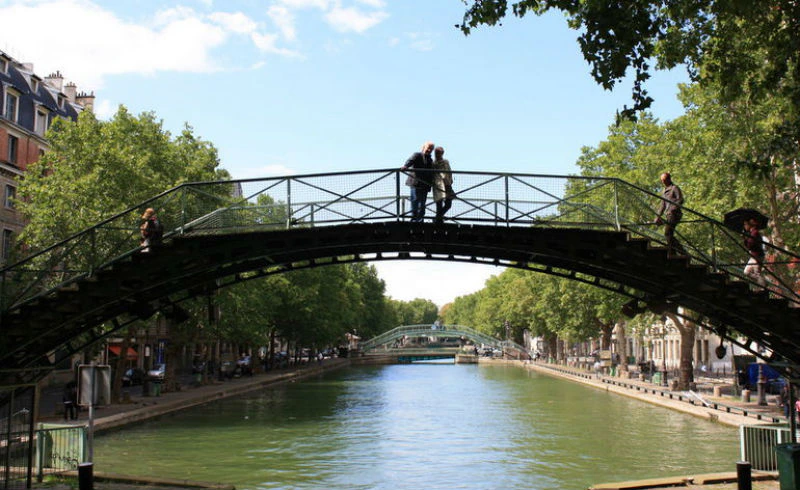 Two people standing on an arched metal bridge over a tree-lined canal with another bridge in the background.