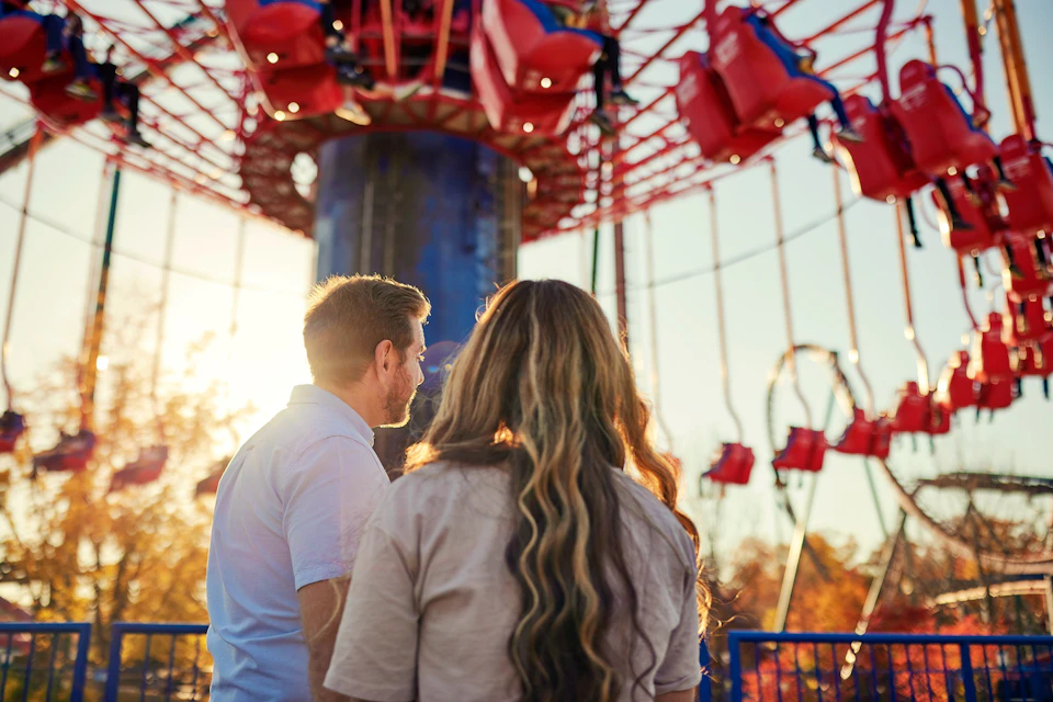 Two people stand in front of a swing ride at an amusement park, with the sun shining in the background.