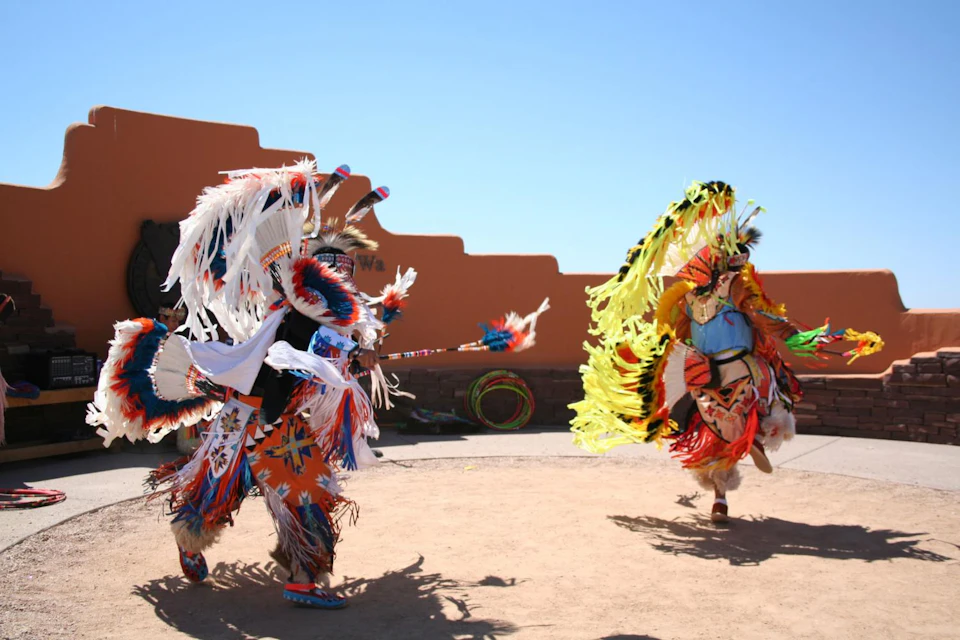 Two individuals in vibrant, traditional attire perform a dance outdoors under a clear blue sky beside an orange adobe wall.