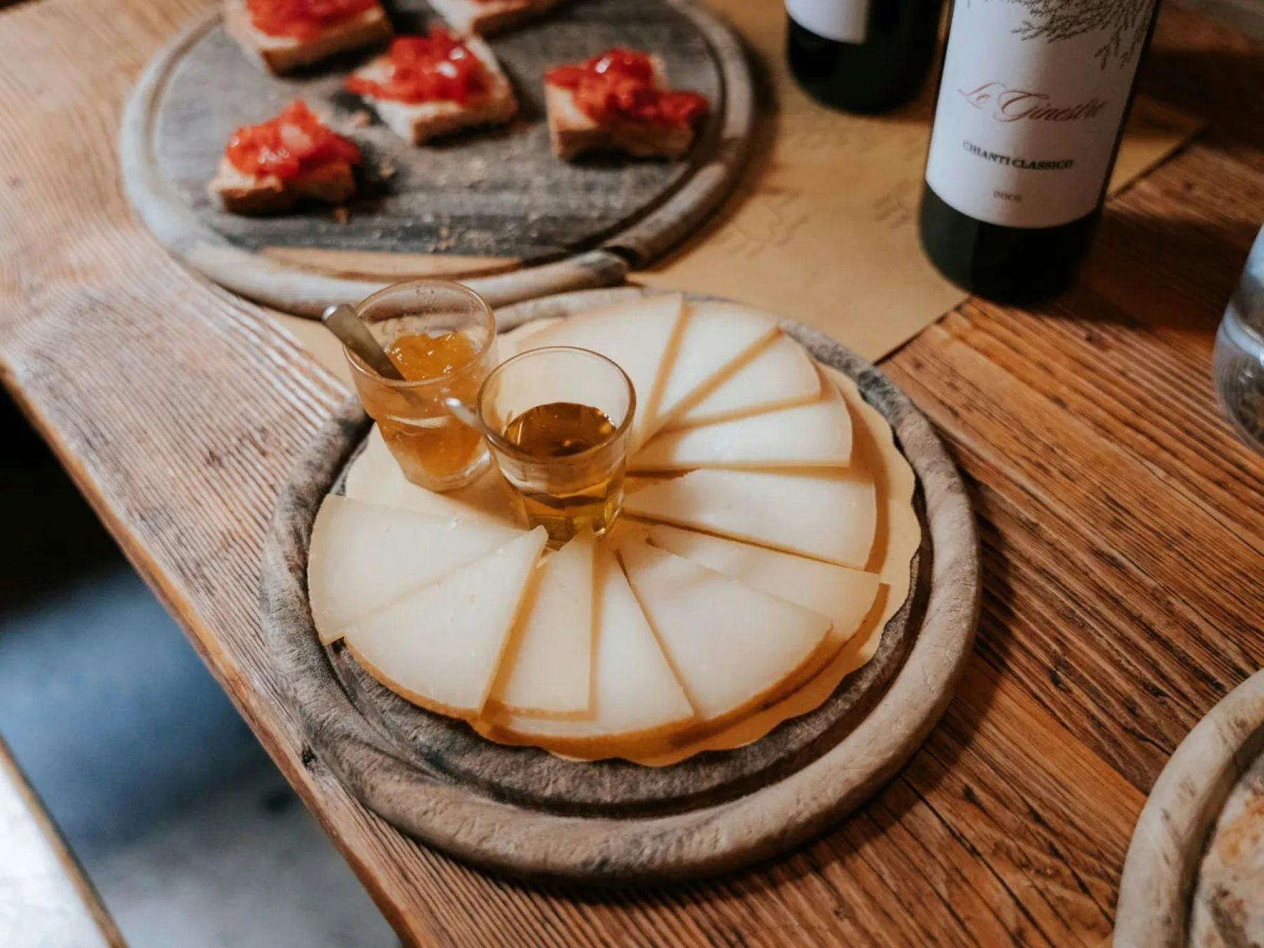 Un vassoio di legno con fette di formaggio e due bicchieri di liquido ambrato, pane con condimenti e bottiglie di vino sullo sfondo.
