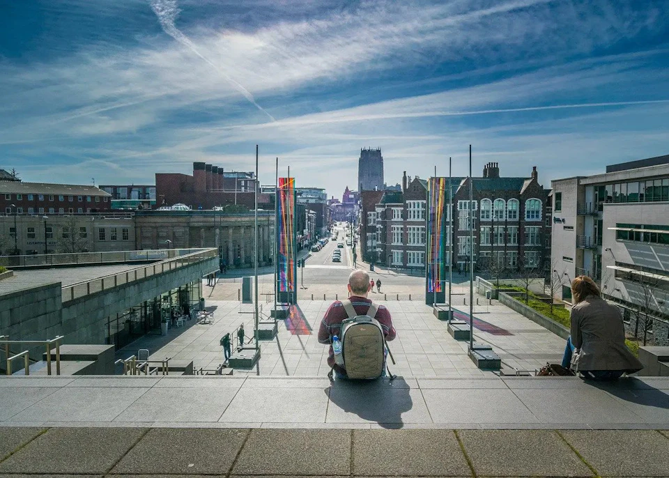 A person with a backpack sits on wide steps overlooking a city street flanked by colorful flags under a blue sky.