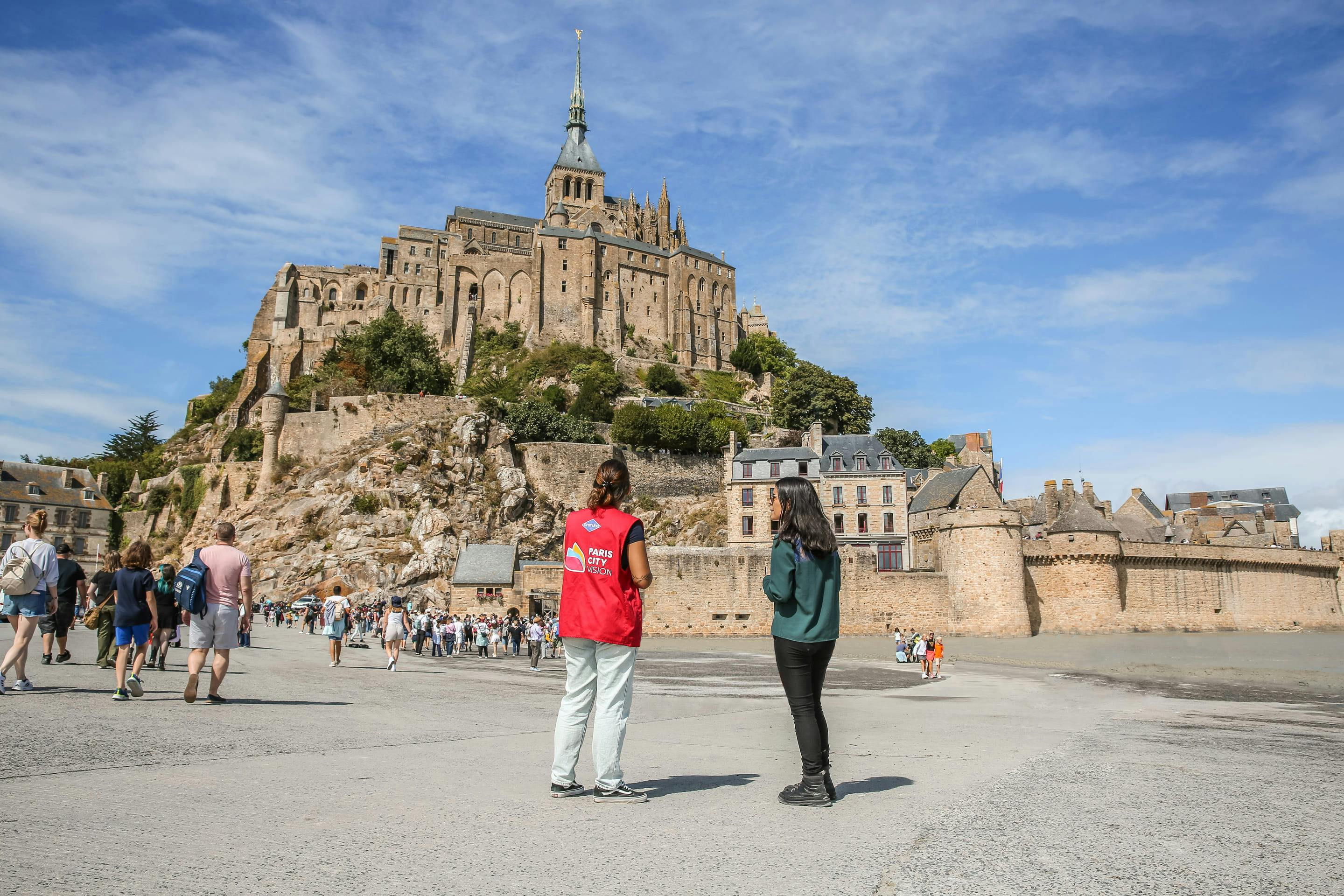 Dva lidé stojí před Mont Saint-Michel pod modrou oblohou. Jeden nosí červenou vestu a v pozadí je vidět turista.