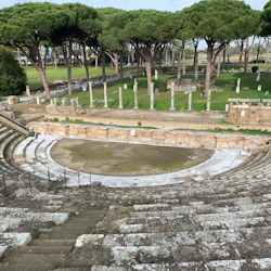Forntida stenamfiteater med halvcirkelformade sittplatser, omgiven av stora träd och ruiner i bakgrunden.