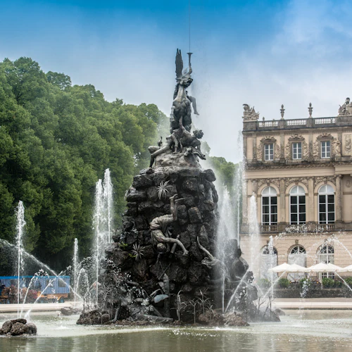 Verschnörkelter Brunnen mit Statuen, Wasserspritzern und einem historischen Gebäude im Hintergrund. Üppige grüne Bäume umgeben die Szene.