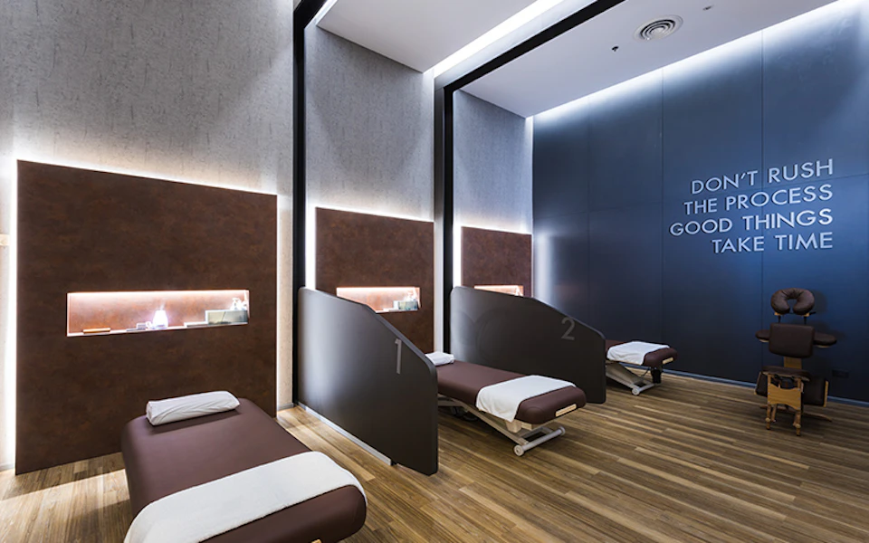 A modern spa room with three reclining beds separated by partitions, featuring sleek lighting and wooden flooring.