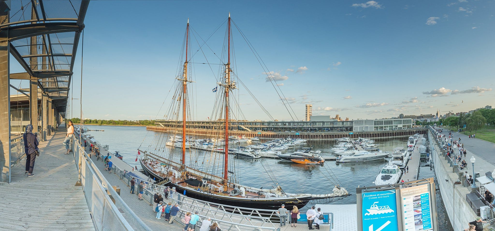 人们在停泊的帆船和游艇旁边的海滨木板路上行走。 后面有一栋大楼。