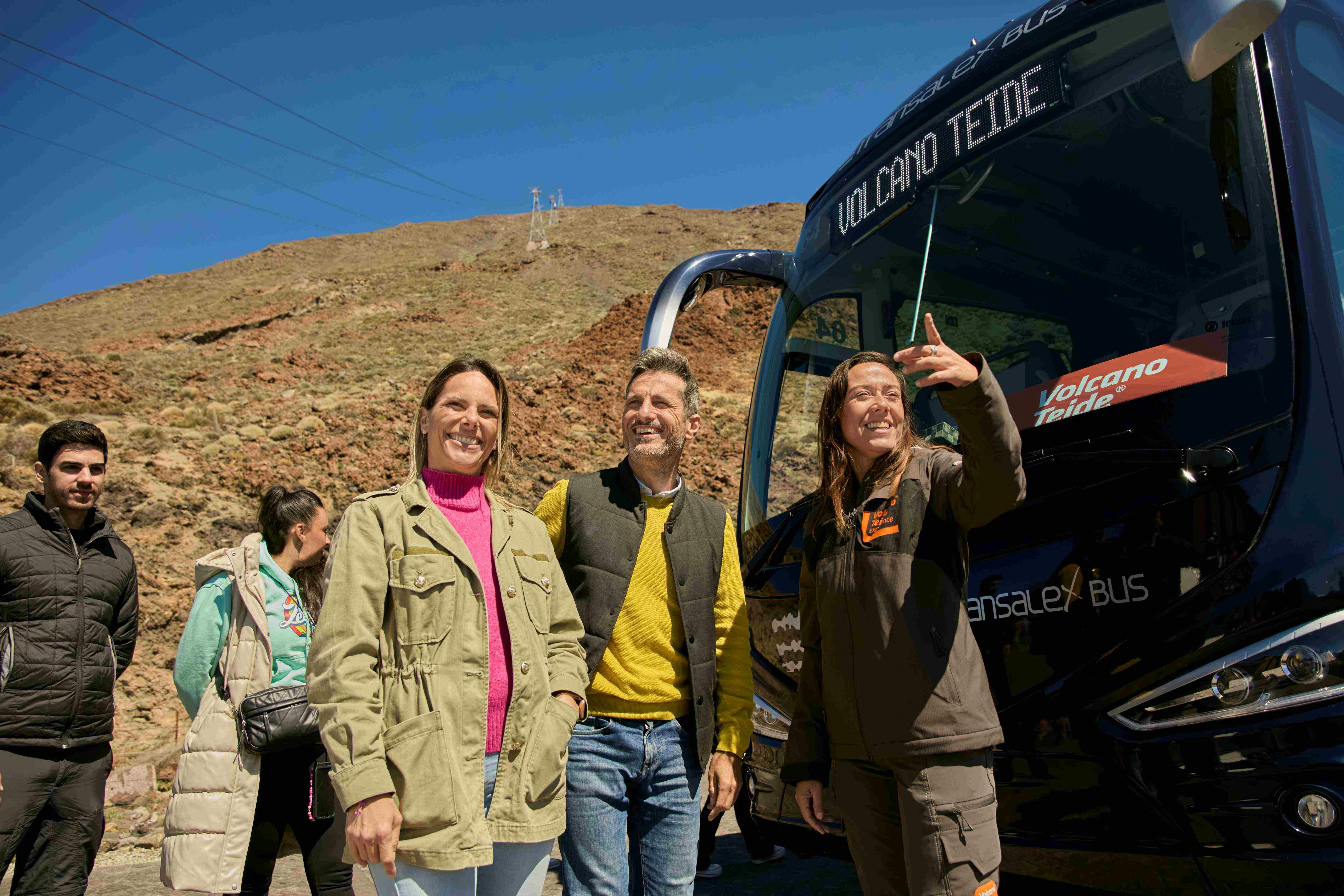 Three people stand smiling next to a bus labeled "Volcano Teide." One person is taking a selfie. Rocky terrain is in the background.