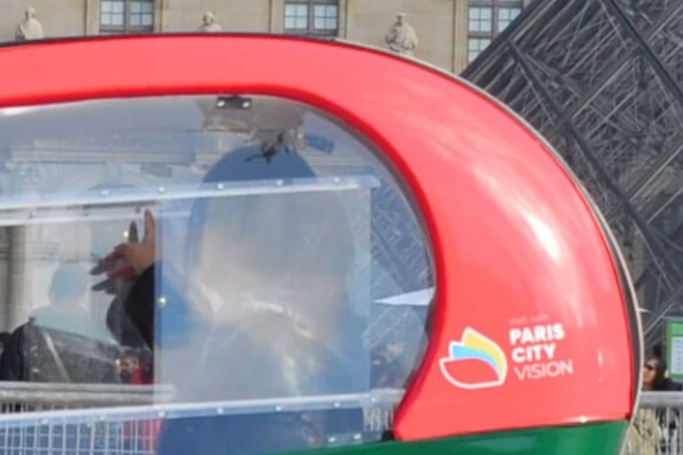 Two green and red pedal-powered taxi vehicles parked in front of the Louvre Pyramid on a sunny day.