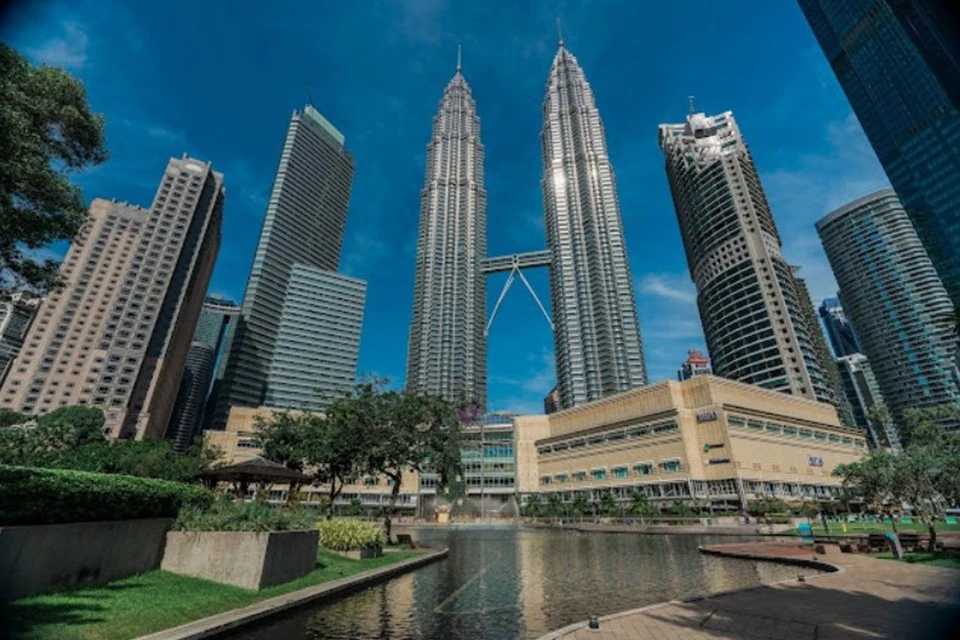 A view of the Petronas Twin Towers surrounded by other buildings, with a park and water feature in the foreground.