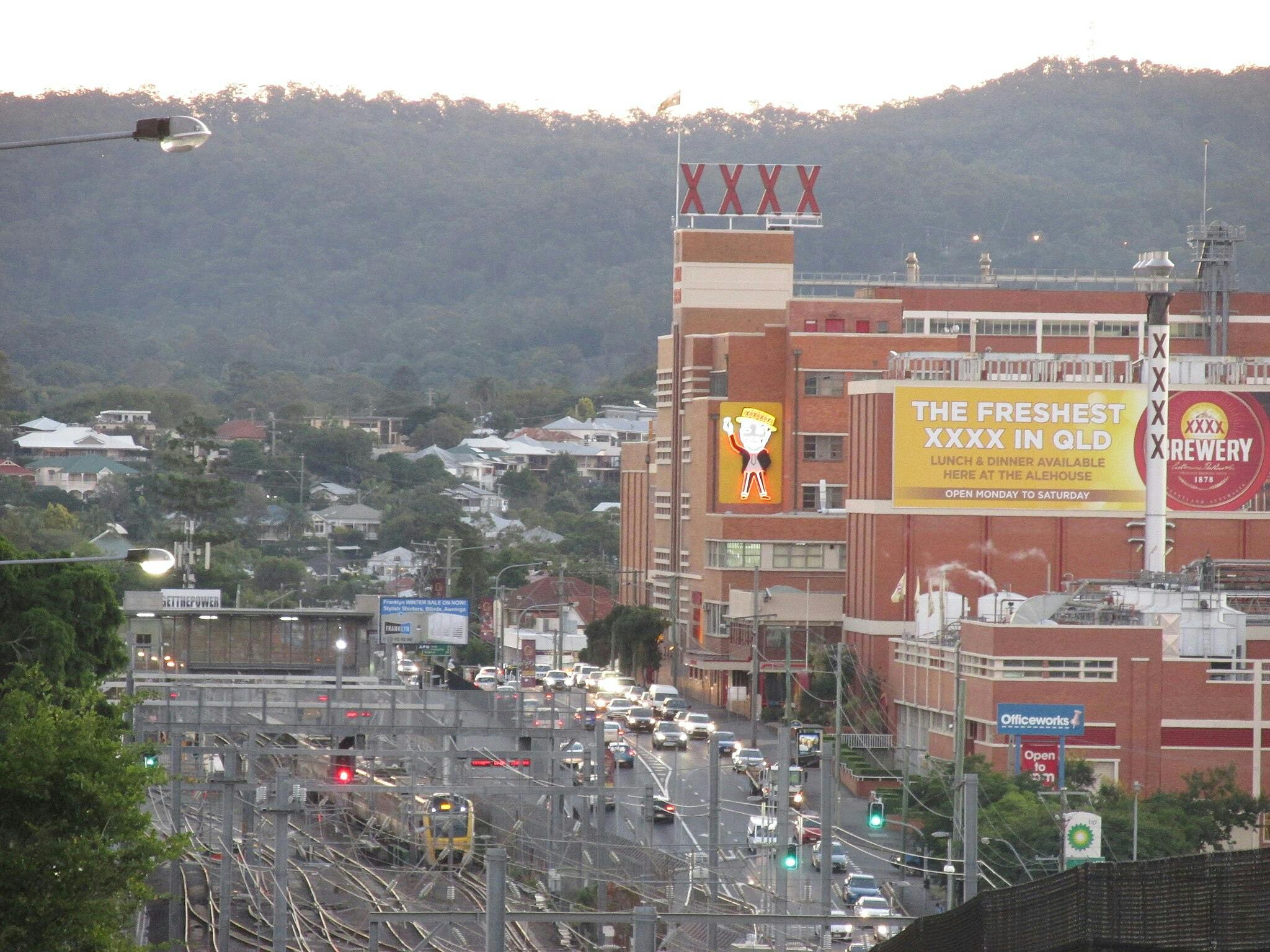 大きな「XXXX」看板があり、鉄道に隣接しており、丘の上には家や緑が広がっています。