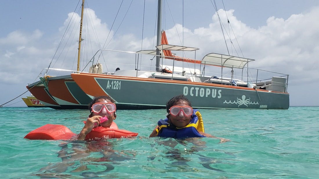 Two children wearing snorkeling gear and life vests swim in clear turquoise water near a sailboat named "Octopus."