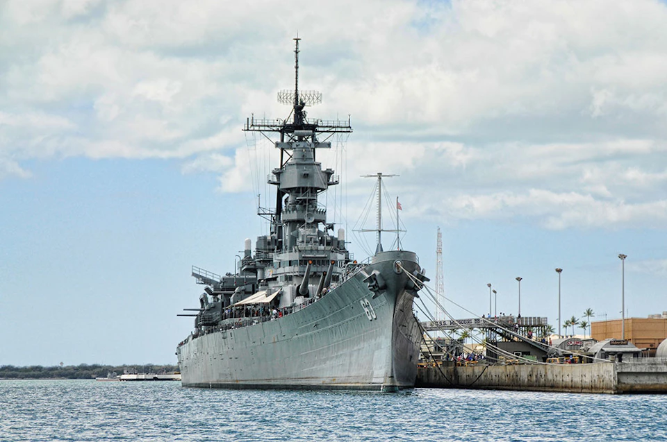 Large gray battleship docked at a port, with visible antennas and turrets, under a cloudy sky.