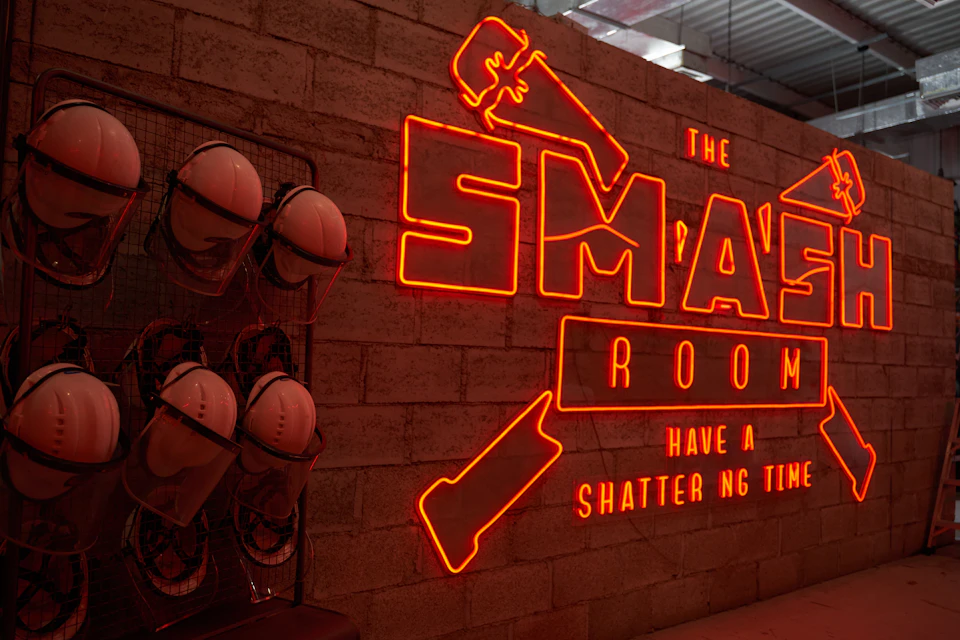Neon sign reading "The Smash Room" with safety helmets on racks against a brick wall. Text includes "Have a Shattering Time."
