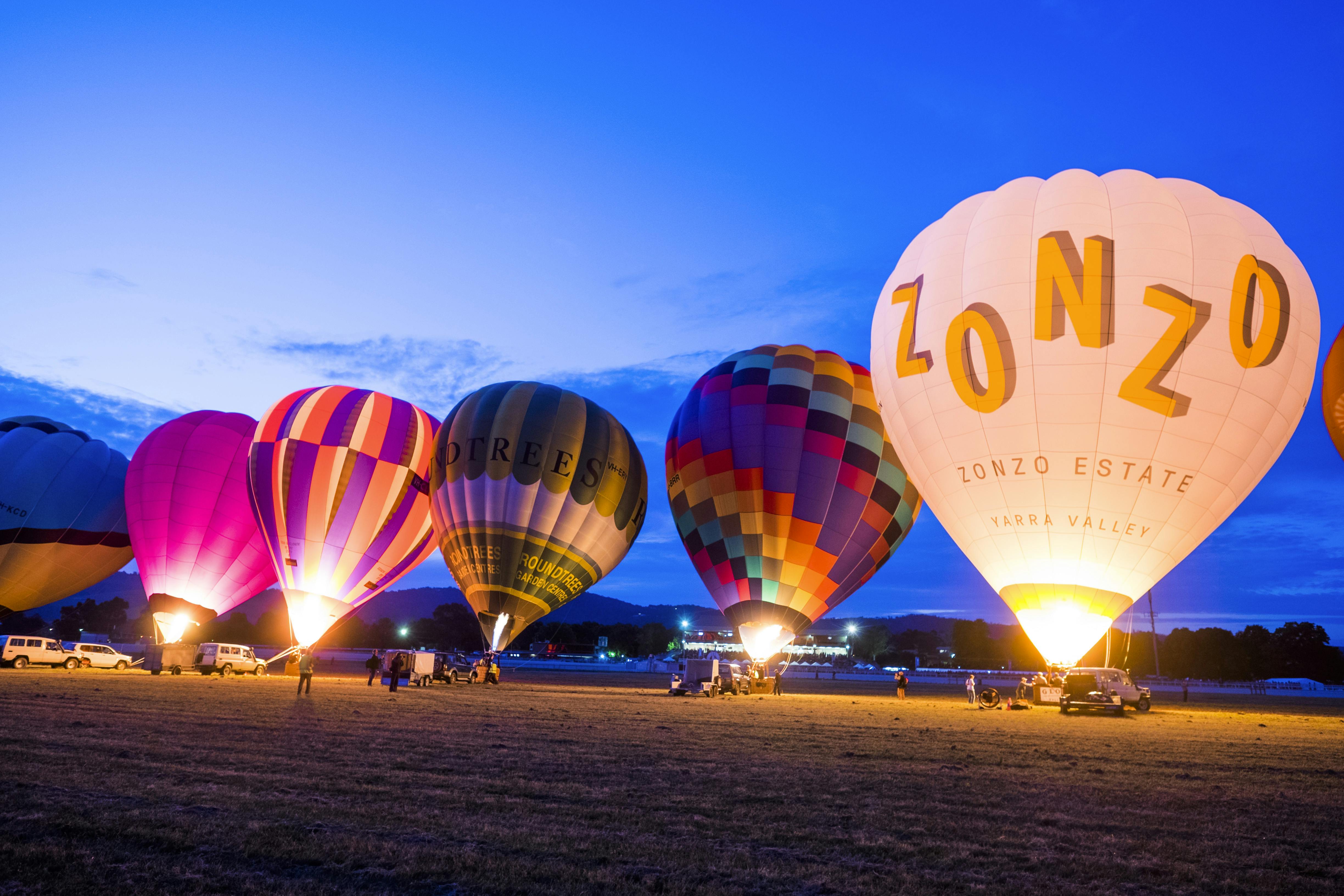 一群五颜六色的热气球在黄昏时分在开阔的田野上充气，周围有几个人和车辆。