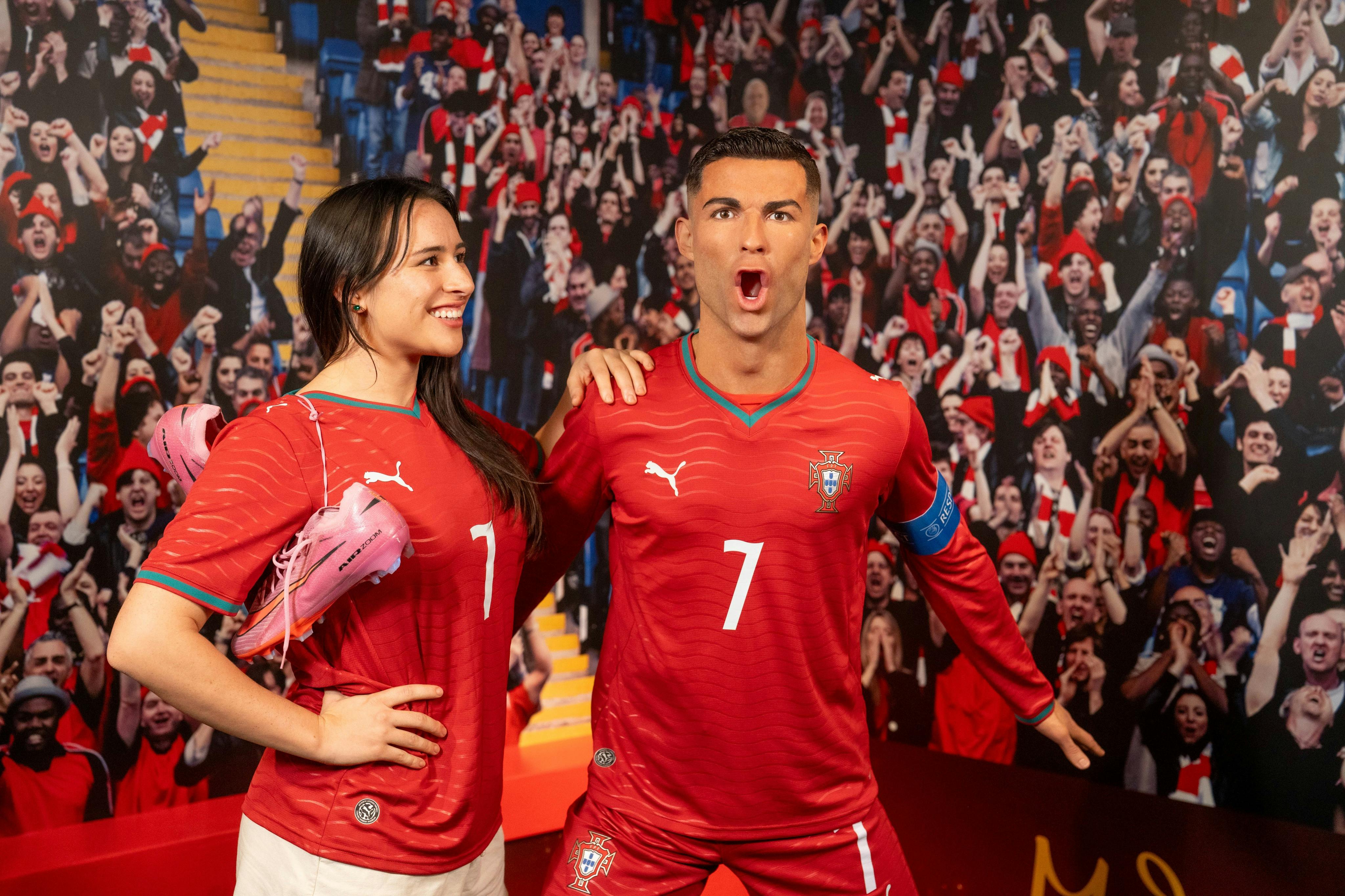 A person poses with a wax figure of a soccer player in a red uniform in front of a cheering crowd backdrop.