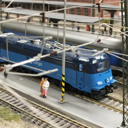 Modelltåg med ett blått lastlok, spår, järnvägsstation, kraftledningar och miniatyrpersoner på plattformen.