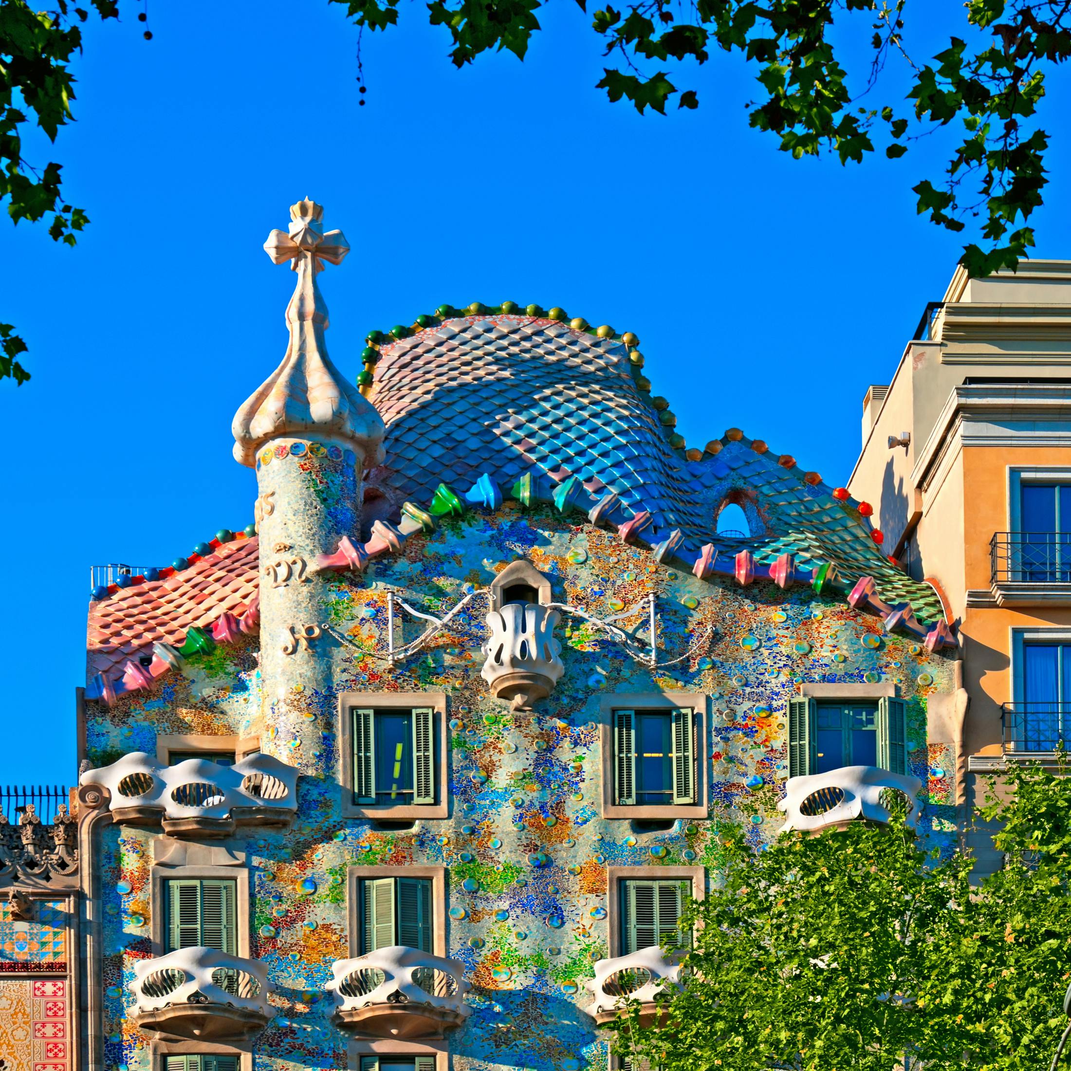 Bâtiment coloré avec des carreaux de mosaïque, une architecture courbée et des fenêtres uniques, sur fond de ciel bleu vif et entouré d'arbres.